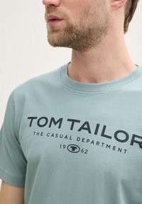 Mėtų spalvos medvilninė marškinėliai su apvalia iškirpte ir juodu užrašu: "TOM TAILOR THE CASUAL DEPARTMENT 1962" paryškintomis raidėmis.