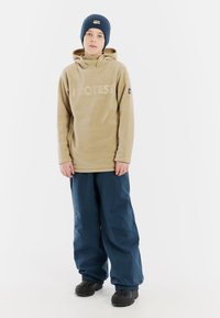 Beige fleece hoodie met "PROTEST" logo, gecombineerd met blauwe waterdichte sneeuwbroek en zwarte winterlaarzen. Lichtgewicht, casual pasvorm.