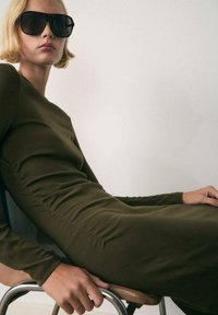 Vestito lungo a maniche lunghe verde oliva scuro con dettagli arricciati, abbinato a occhiali da sole neri oversize. Modella seduta su una sedia contro uno sfondo neutro.