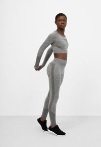 Sivý dlhý rukáv crop top a legíny s vysokým pásom s textúrovanými akcentmi. V kombinácii s čiernymi atletickými topánkami, na neutrálnom pozadí.