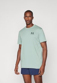 Világoszöld pamut póló, kerek nyakkivágással és rövid ujjakkal, kis fekete logóval a mellkason. Tengerészkék sorttal viselve.