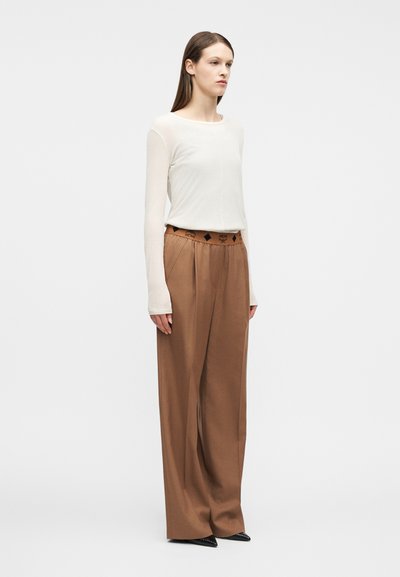 Jeune femme portant un haut à manches longues crème et un pantalon large marron taille haute, avec des chaussures pointues noires, debout de profil.