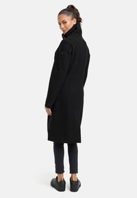 Manteau en laine noir avec col montant, manches longues et coupe ajustée. Doté de poches latérales et d'un détail de couture notable. Accompagné de baskets noires.