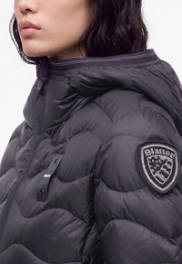 Femme portant une veste matelassée noire avec un col montant et un patch de marque sur le haut du bras.
