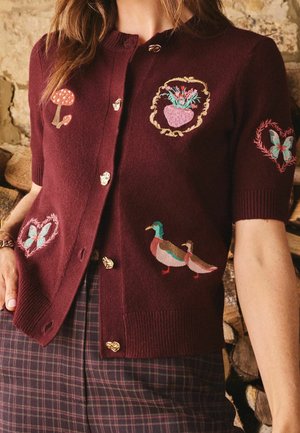 Frau trägt einen kastanienbraunen Kurzarm-Cardigan mit bestickten Pilzen, Blumen, Enten und Schmetterlingen, kombiniert mit karierten Hosen.