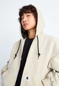 Beige zip-opp hettegenser med svart foret hette, snorer og frontlommer. Har et logo-patch på venstre lomme. Svart T-skjorte under.