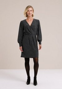 Čierne obopínajúce šaty s dlhými, nafúknutými rukávmi, vyrobené z trblietavého materiálu. Majú hlboký výstrih do V a priliehavý pás, skombinované s čiernymi pančuchami a podpätkami.