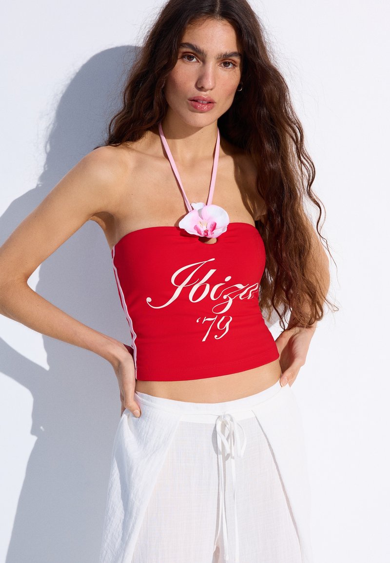 Femme aux longs cheveux bruns portant un haut rouge bustier avec l'inscription « Ibiza '79 » et un pantalon blanc à cordon, accessoirisé d'un collier de fleurs roses.