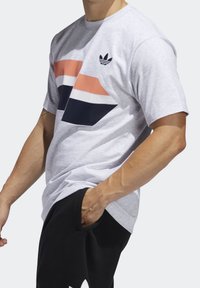 Šedé tričko s krátkým rukávem s geometrickou grafikou v oranžové, bílé a námořnické modři. Logo v levém horním rohu. Bavlněný materiál, volný střih.