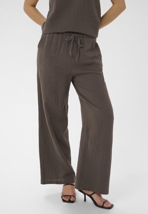 Femme portant un pantalon ample texturé marron foncé avec taille à cordon et sandales noires à talons hauts, debout les mains dans les poches.