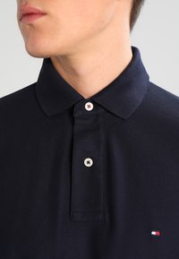 Námornícky polo tričko s texturovanou tkaninou, dva gombíky s kontrastnými akcentmi a malým logom v červenej, bielej a modrej na spodnej strane.