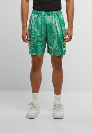 Man wearing green patterned shorts with yellow and white patches, white socks, white sneakers, and a black shirt, standing on gray floor.