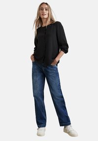 Camicia nera con bottoni, in tessuto strutturato, maniche lunghe e vestibilità rilassata, abbinata a jeans a gamba larga blu e sneakers bianche.