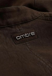 Hnědá texturovaná látka s viditelným švem. Obsahuje kovovou štítek "ombre", našitou na tmavší části.
