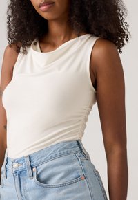 Frau in einem ärmellosen weißen, gerafften Oberteil und hellblauen, hochsit Sitzen Jeans, mit lockigem Haar, das über die Schultern sichtbar ist.