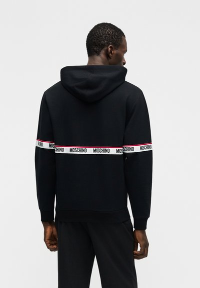 Άνδρας με πλάτη φοράει μαύρο φούτερ με κουκούλα με λευκή ταινία και το κείμενο "MOSCHINO" γύρω από το στήθος και τα χέρια.