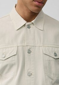 Licht beige denimjacket met een klassiek kraag, twee borstzakken en zilveren knopen. Heeft een gestructureerde stof en zichtbare stiksels.