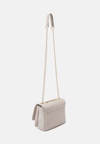 Kremowa pikowana torebka z tłoczonym wzorem, zamknięciem na klapę i złotym łańcuszkowym paskiem z skórzaną poduszką na ramię, wisząca na białym tle.