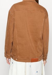 Veste en denim brun à coupe décontractée, avec des poignets à boutons, des détails de couture et une patch en cuir marquée au dos de l'ourlet.