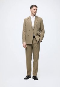 Beiges, schmal geschnittenes, doppelt geknöpftes Sakko mit Einschultern, seitlichen Taschen und schwarzen Dressschuhen. Darunter ein weißes Hemd mit Kragen.