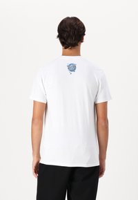 Camiseta de algodón blanca con un pequeño gráfico azul en la parte posterior, que muestra el texto "SANTA CRUZ" rodeado de elementos decorativos.