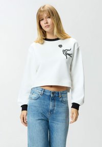 Felpa bianca corta con dettagli neri, caratterizzata da un ricamo di un uccello e un cuore. Abbinata a jeans di denim azzurro chiaro.