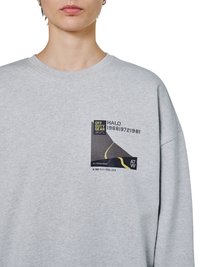 Osoba nosząca jasnoszarą bluzę z małą czarno-żółtą grafiką i nadrukowanym tekstem na piersi.