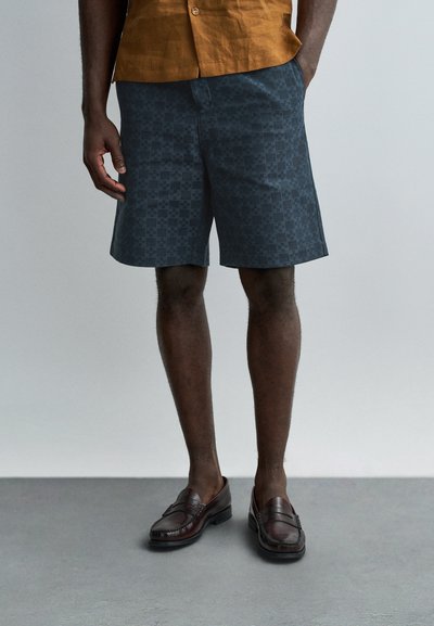 Homme portant un short sombre à motifs, des mocassins marron et une chemise boutonnée orange-brun, les mains dans les poches, debout sur un sol gris.