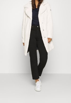 Femme portant un manteau en fausse fourrure blanche, un pantalon noir taille haute, un top bleu marine et des baskets blanches, debout devant un fond blanc uni.