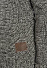 Detalle de un suéter gris de punto que presenta una etiqueta de cuero con la marca "Solid". La textura es gruesa y acanalada, mostrando un diseño acogedor.