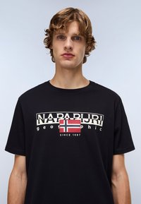 Tricou negru din bumbac, cu un design grafic ce include textul "NAPAPIJRI" și un patch al steagului Norvegiei. Tricoul are un guler rotund și mâneci scurte.