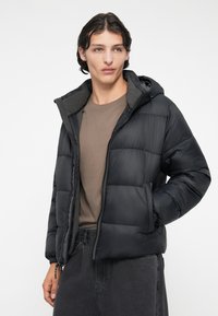 Chaqueta acolchada negra con capucha, con cierre de cremallera, puños elásticos y un diseño acolchado. Lleva puesta sobre una camiseta marrón y pantalones grises.