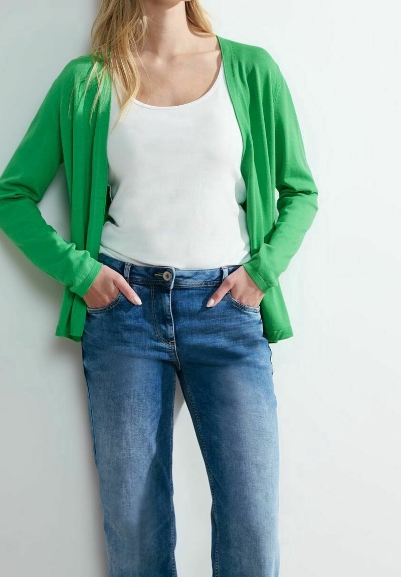 Cardigan verde sopra una canotta bianca, abbinato a jeans blu denim. Il cardigan ha una vestibilità comoda con maniche lunghe.
