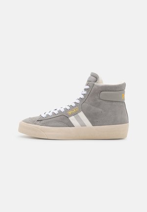Szary sneaker za kostkę z białymi sznurowadłami, dwoma białymi paskami po bokach, beżową podeszwą i metką „Polo” przy sznurowadłach na jednolitym tle.