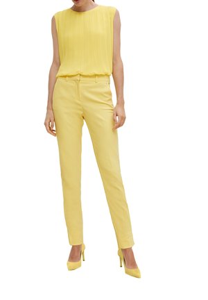 Vrouw draagt een mouwloze geplooide gele top, strakke gele broek en gele hoge hakken, staand tegen een witte achtergrond.