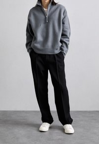 Pull gris clair à col zippé avec poignets et ourlet côtelés, associé à un pantalon plissé noir et des baskets blanches, debout devant un fond neutre.