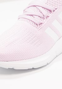 Světle růžový pletený teniskový boty s bílou podrážkou, tkaničkami a třemi vertikálními bílými pruhy na boku, zobrazené v detailním záběru.