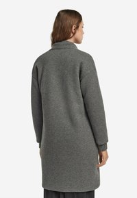 Lang, grå uldfrakke med høj krave, sænkede skuldre og ribbede ærmeafslutninger. Glat tekstur, mellemlang design, minimale syninger detaljer.