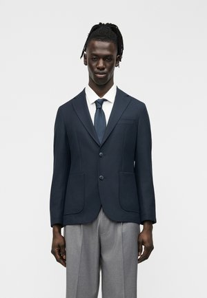 Άνδρας με ντραντλοκς που φοράει μπλε μπλέιζερ, λευκό πουκάμισο, σκούρο γραβάτα και γκρι παντελόνι, στέκεται μπροστά σε απλό γκρι φόντο.