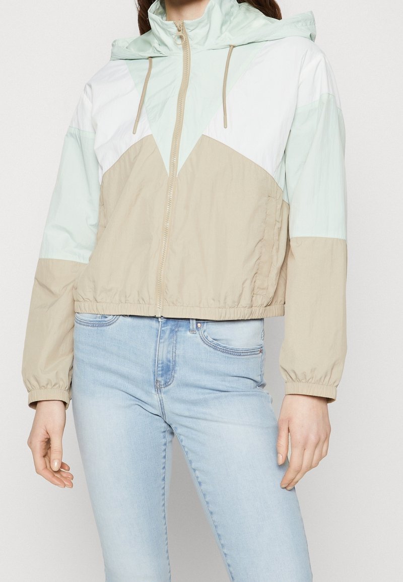 Kurtka przeciwwiatrowa w blokach kolorów o miętowo-zielonym i beżowym wzorze. Posiada kaptur, zamek błyskawiczny, elastyczne mankiety oraz krótki krój.