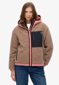 Veste en polaire marron avec une doublure rose, des accents marins et une fermeture éclair à l'avant. Elle est dotée d'une capuche et d'une poche sur la poitrine, conçue pour la chaleur et le confort.