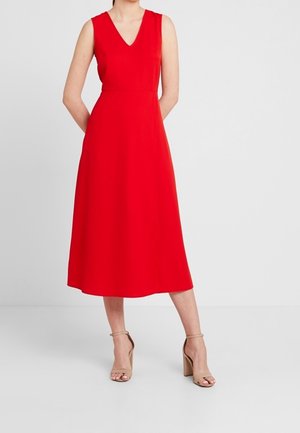 Kobieta ubrana w czerwoną sukienkę midi bez rękawów z dekoltem w serek oraz beżowe sandały na obcasie z paskiem wokół kostki, stojąca na tle jednolitego białego tła.