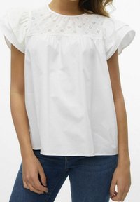 Donna che indossa una blusa bianca leggera con dettagli floreali ricamati sullo scollo e maniche a sbuffo, abbinata a jeans blu.