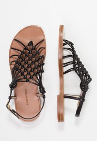 Sandales en cuir tressé noir avec une semelle plate, dotées d'une semelle intérieure marron et d'une bride de cheville réglable. Le design détaillé met en valeur la texture et la forme.