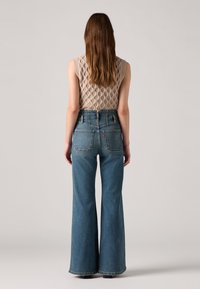 Jeans azuis de cintura alta e boca de sino, com um design clássico de cinco bolsos e costura em contraste. Top sem mangas bege tricotado com um padrão texturizado.