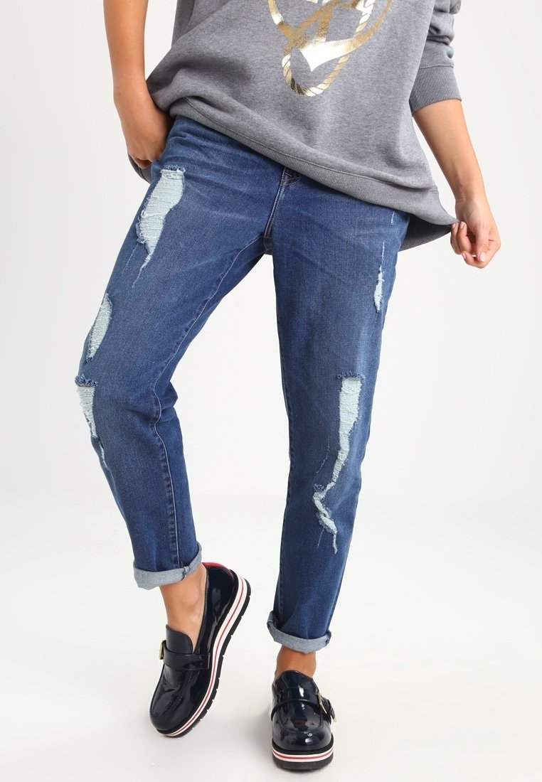 Vihreät siniset pellavat farkut, joissa on useita repeämiä, kapeneva istuvuus, rullatut hihansuut ja keskipesun rakenne. Yhdistetty tummiin kiiltäviin kenkiin.