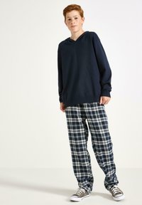 Ein rothaariger Junge steht mit einer dunkelblauen Kapuzenjacke, dunkelblau-weiß karierten Hosen und grauen Turnschuhen vor einem weißen Hintergrund.
