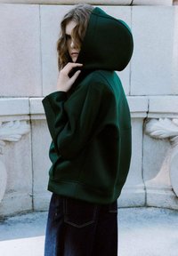 Sudadera con capucha de color verde oscuro, con una capucha grande, textura suave y corte holgado, combinada con unos vaqueros azul oscuro. Diseño minimalista, sin logotipos visibles.