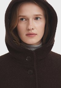 Manteau à capuche texturé marron avec deux boutons noirs, porté sur un haut gris à col montant, mettant en avant son design confortable et son matériau doux.