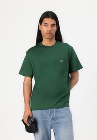 T-shirt in cotone verde con scollo rotondo e maniche corte, caratterizzato da una piccola tasca e un logo sul petto. Indossato con jeans blu.